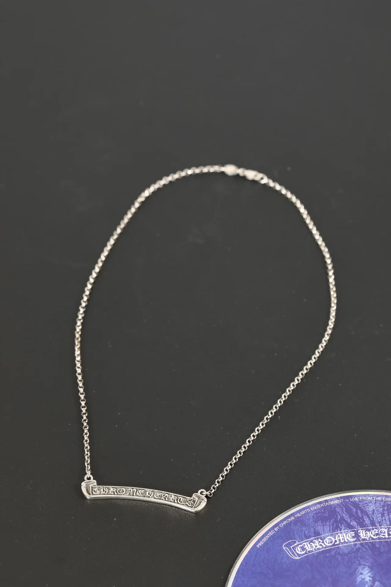 Серебряное колье Chrome Hearts с планкой Scroll Logo на тёмном фоне