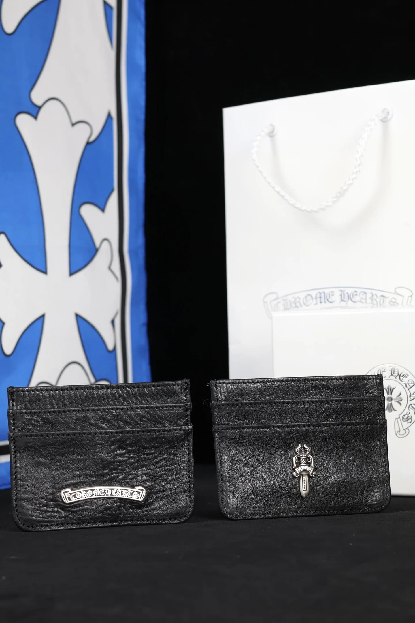 Два чёрных кожаных картхолдера Chrome Hearts с серебряной фурнитурой на фоне фирменного пакета