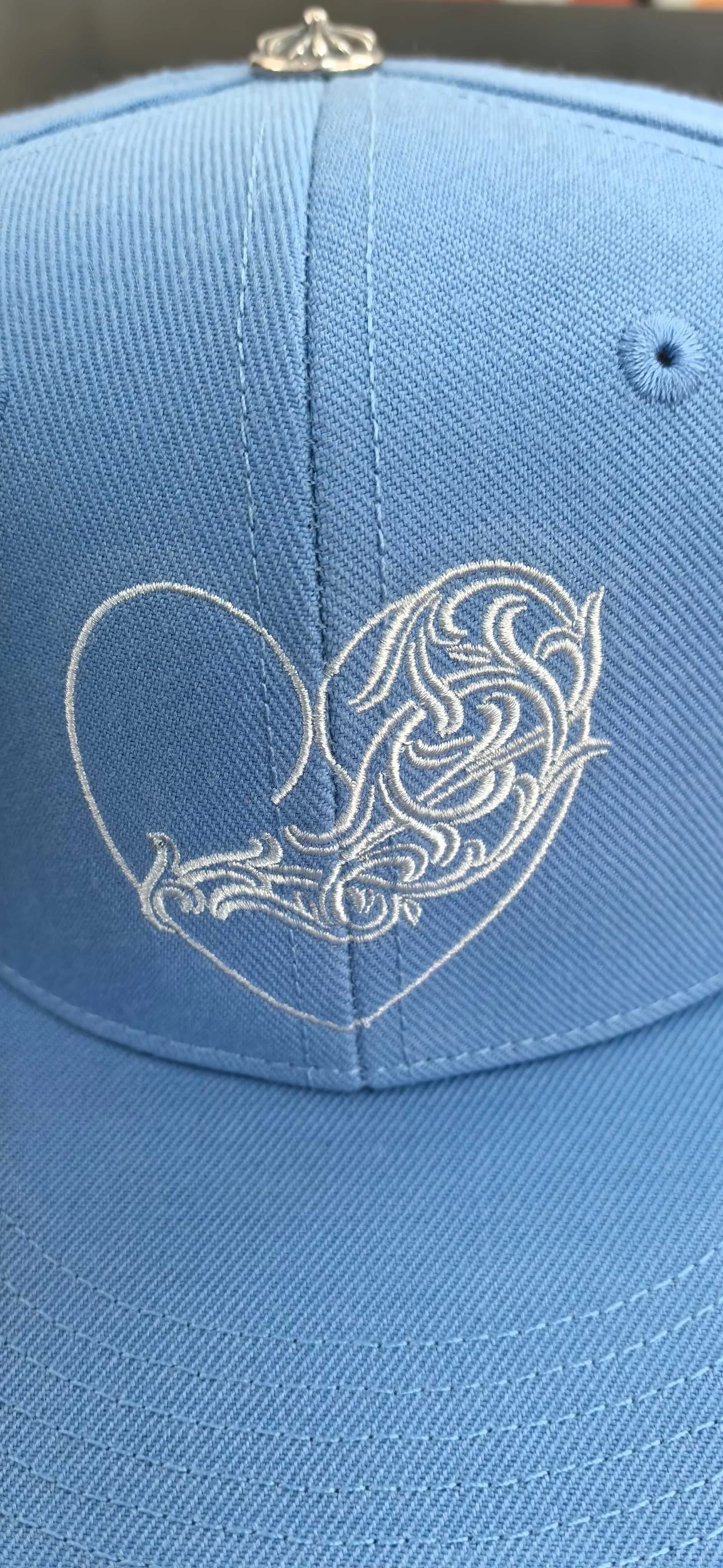 Крупный план вышивки в виде белого сердца на голубой бейсболке Chrome Hearts